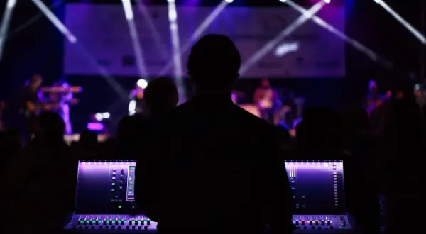 Silueta de técnico de sonido operando una mesa de mezclas en un plano de escenario musical, con luces púrpuras y blancas iluminando la banda al fondo.
