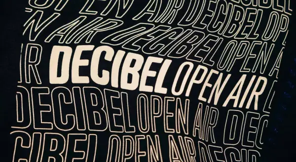 Texto en una camiseta negra con el lema "Decibel Open Air" repetido en letras blancas onduladas, evocando la energía de un festival.