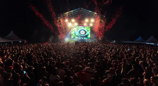 Multitud en un concierto al aire libre, con confeti rojo cayendo sobre el escenario y una pantalla gigante. El **bass** retumba en el parque.