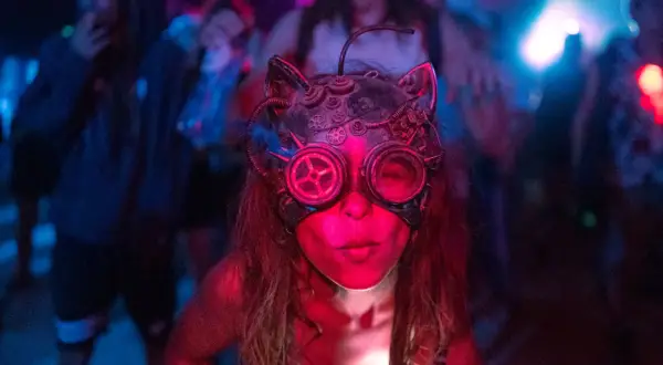 Una persona con una máscara de gato steampunk y gafas de aviador, rodeada de luces de neón y gente bailando. Así es que es la rave.