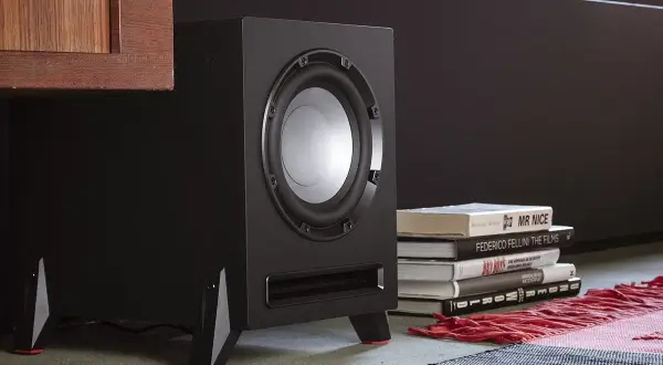 Subwoofer negro con detalles rojos, junto a libros y una alfombra.