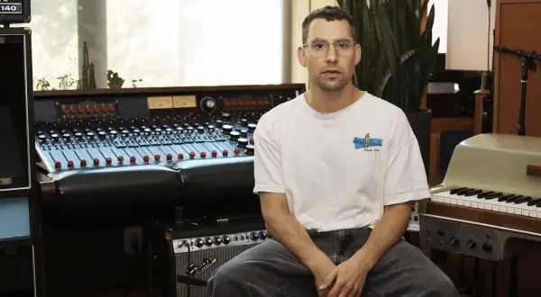 Productor musical con gafas sentado frente a una mesa de mezclas y un teclado en un estudio de grabación.