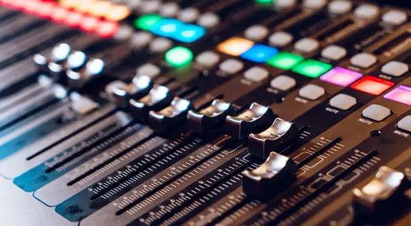 Primer plano de una mesa de mezclas con faders y botones iluminados. La **técnica de sonido** se manifiesta en este panel de control para crear la atmósfera perfecta.