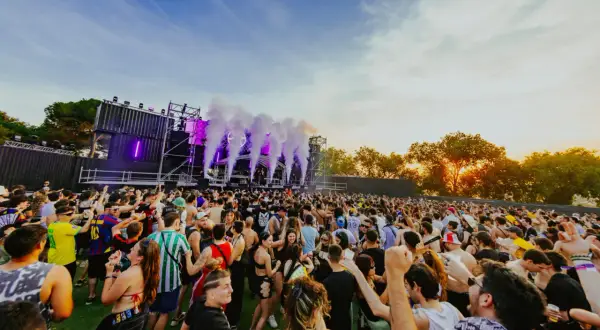 Multitud bailando en un festival al atardecer, con humo y luces moradas saliendo del escenario. ¡Un ambiente genial para los parkineos de donde es!