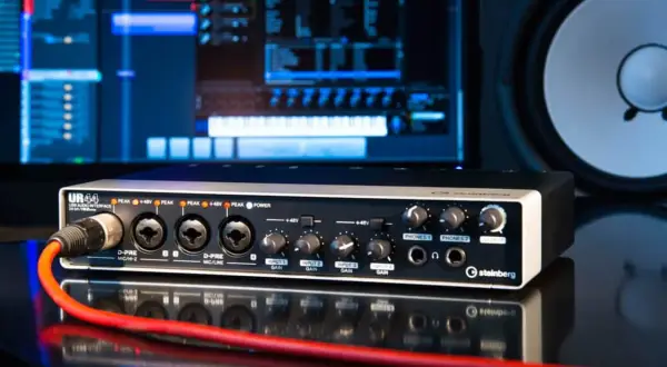 Interfaz de audio Steinberg UR44 con cable rojo conectado, monitor de computadora y altavoz al fondo.