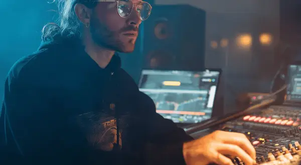 Hombre con gafas y coleta trabajando en un estudio de sonido, creando música electrónica para su blog.