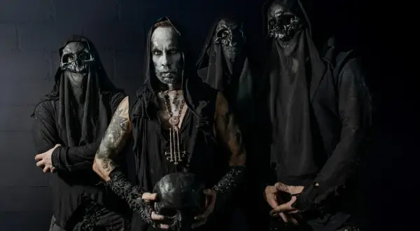 Cuatro figuras encapuchadas, con máscaras de calavera y maquillaje facial pálido, posan sombríamente. El líder sostiene un cráneo, evocando la estética de grupos black metal.