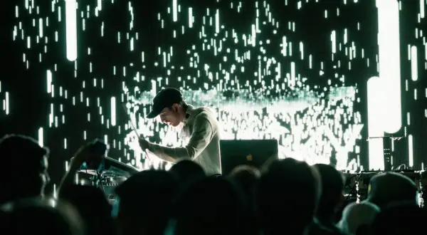 Un DJ techno en acción, con una pantalla de luces verdes y blancas parpadeantes detrás.