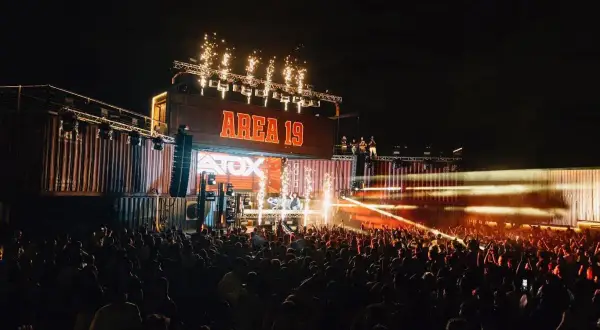 Una multitud celebra en un concierto nocturno, con fuegos artificiales y luces láser iluminando el escenario "AREA 19" y el **nexus fabrik** de contenedores.
