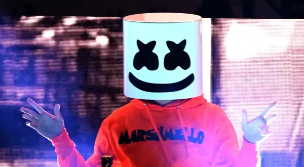 Christopher Comstock, conocido como Marshmello, con su icónica máscara blanca y sudadera naranja, saluda al público en un concierto.
