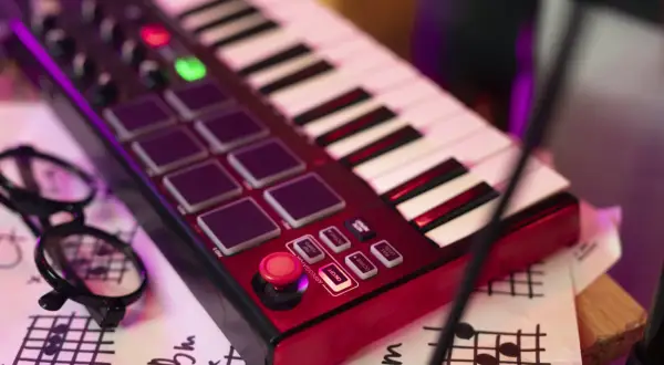 Teclado rojo con pads, botones y teclas blancas y negras. Unas gafas negras y partituras debajo. Es un controlador MIDI para crear música.