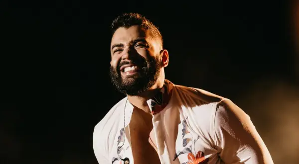 Un cantante brasileño famoso joven ríe con alegría en el escenario, con una camisa blanca estampada y un collar.