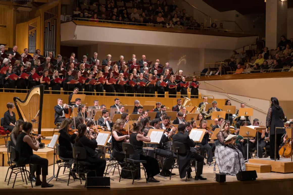 Un coro y orquesta interpretan música en un auditorio. Los cantantes franceses, vestidos de negro, sostienen partituras rojas.