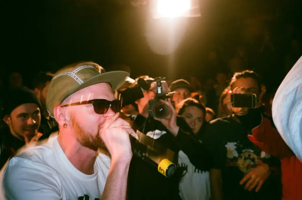 Un rapero con gorra y gafas de sol canta al micrófono, rodeado de público y cámaras. Capturando la energía de los estilos de música urbana.