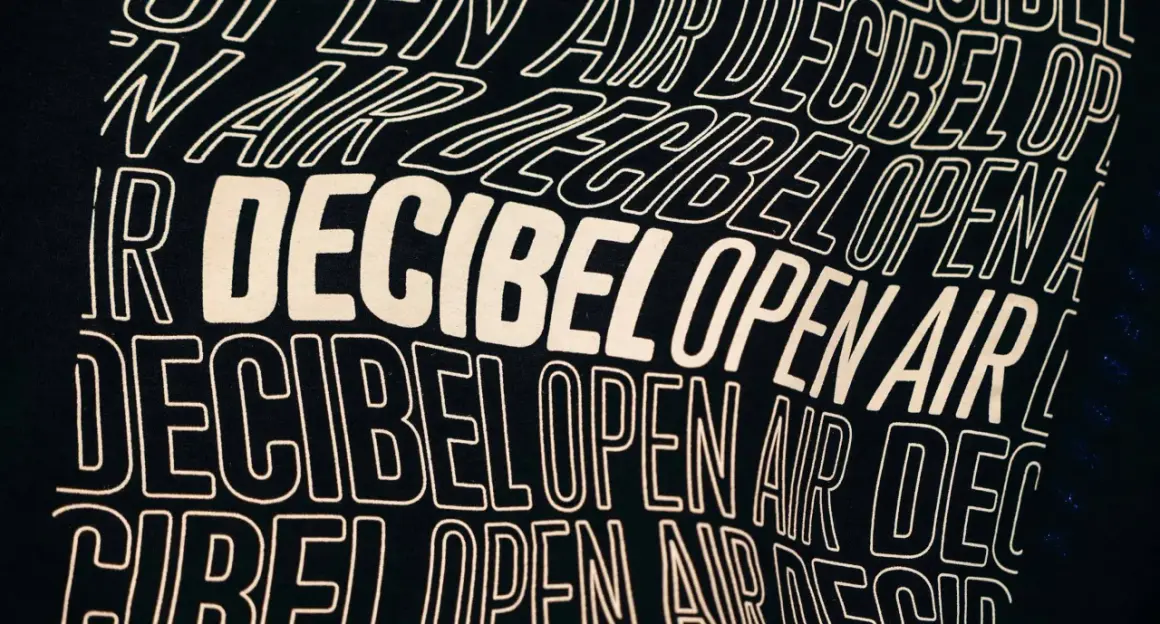 Texto en una camiseta negra con el lema "Decibel Open Air" repetido en letras blancas onduladas, evocando la energía de un festival.