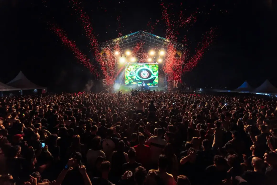 Multitud en un concierto al aire libre, con confeti rojo cayendo sobre el escenario y una pantalla gigante. El **bass** retumba en el parque.