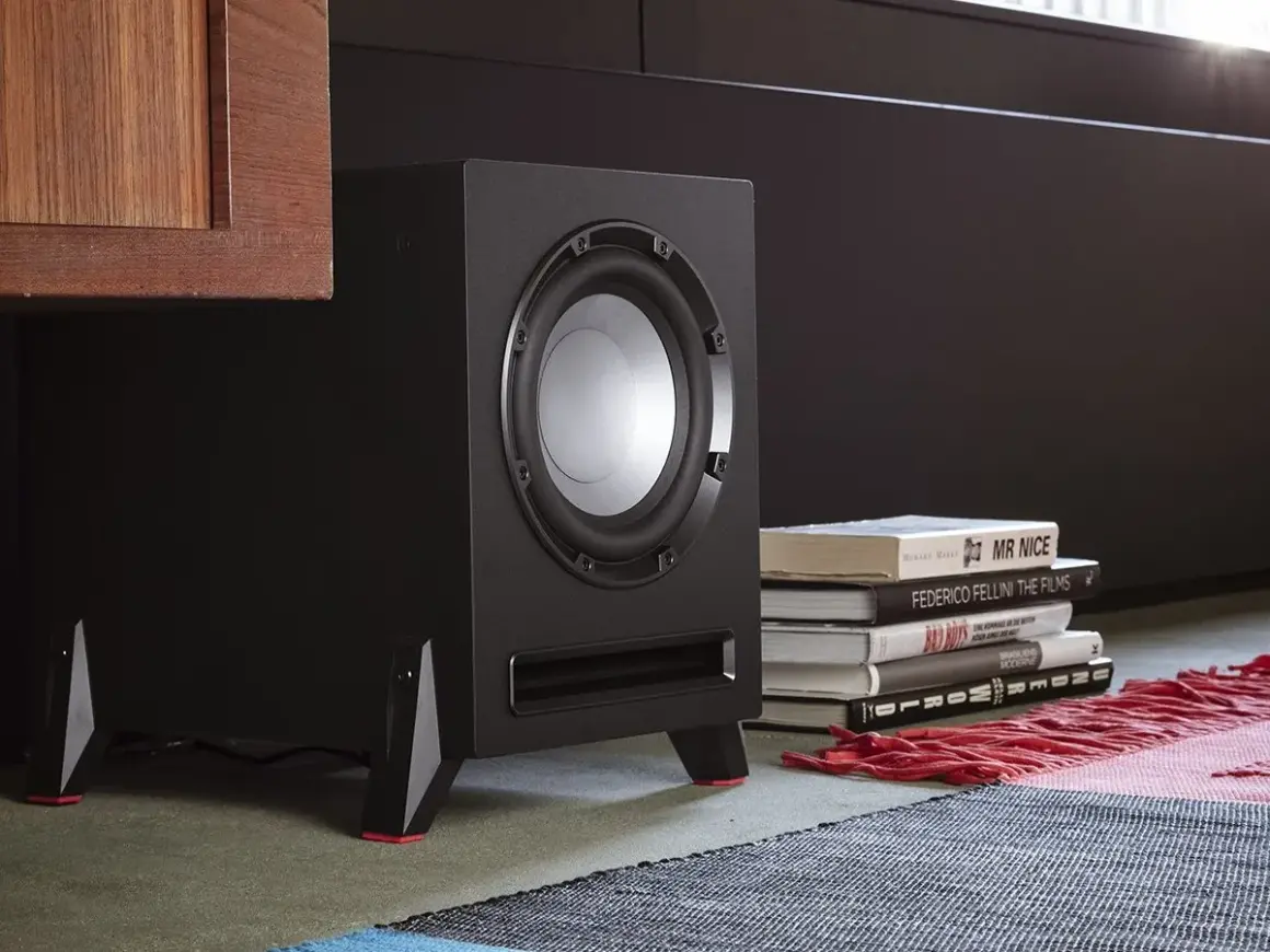 Subwoofer negro con detalles rojos, junto a libros y una alfombra.