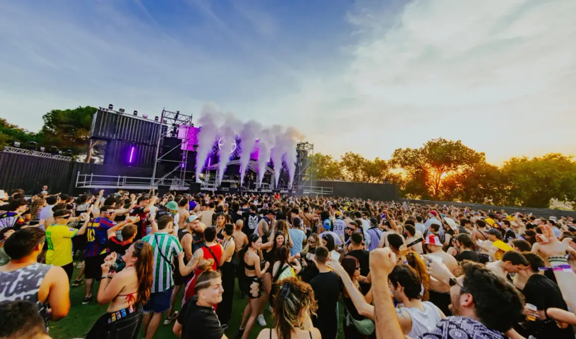 Multitud bailando en un festival al atardecer, con humo y luces moradas saliendo del escenario. ¡Un ambiente genial para los parkineos de donde es!