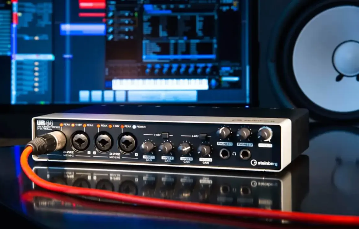 Interfaz de audio Steinberg UR44 con cable rojo conectado, monitor de computadora y altavoz al fondo.
