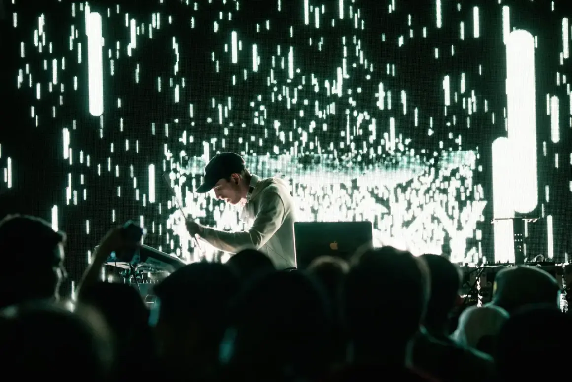 Un DJ techno en acción, con una pantalla de luces verdes y blancas parpadeantes detrás.