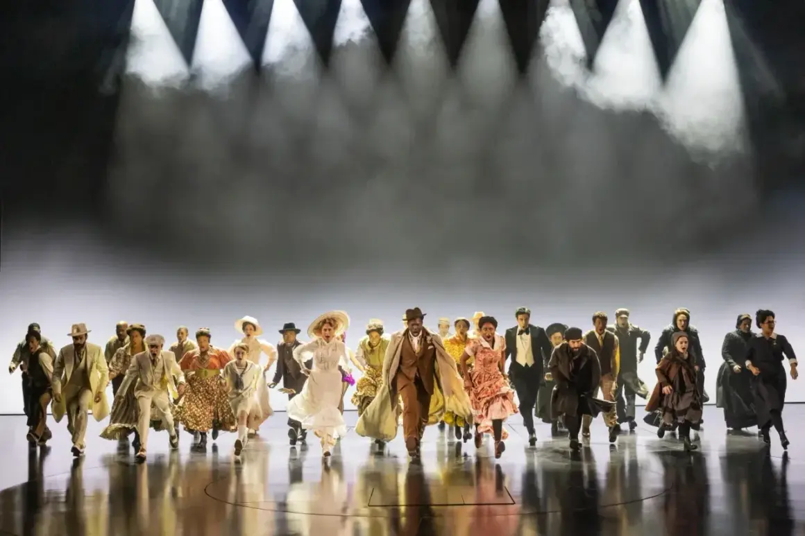 Un grupo diverso de personas corre hacia adelante en un escenario, evocando la energía y el espíritu de "Ragtime, que es".