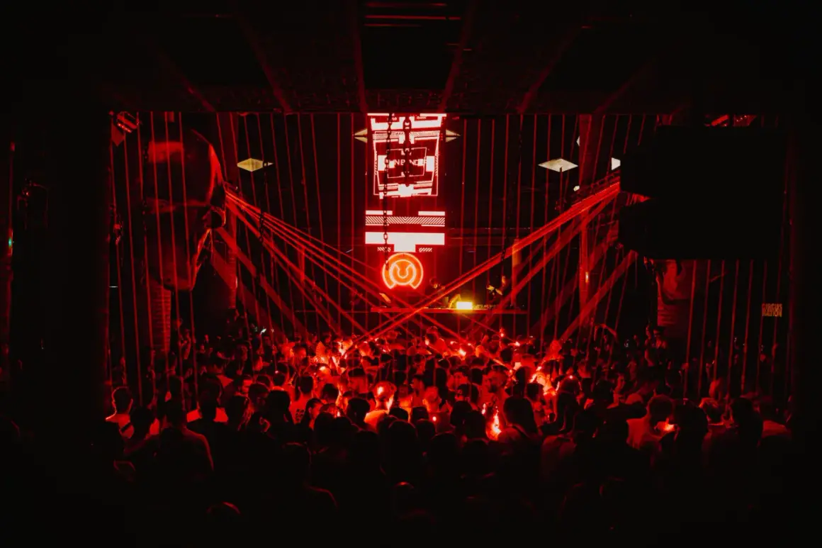 Multitud vibrante en un club nocturno, bañada en luces rojas. El logo de "Circus Nation" ilumina el escenario.