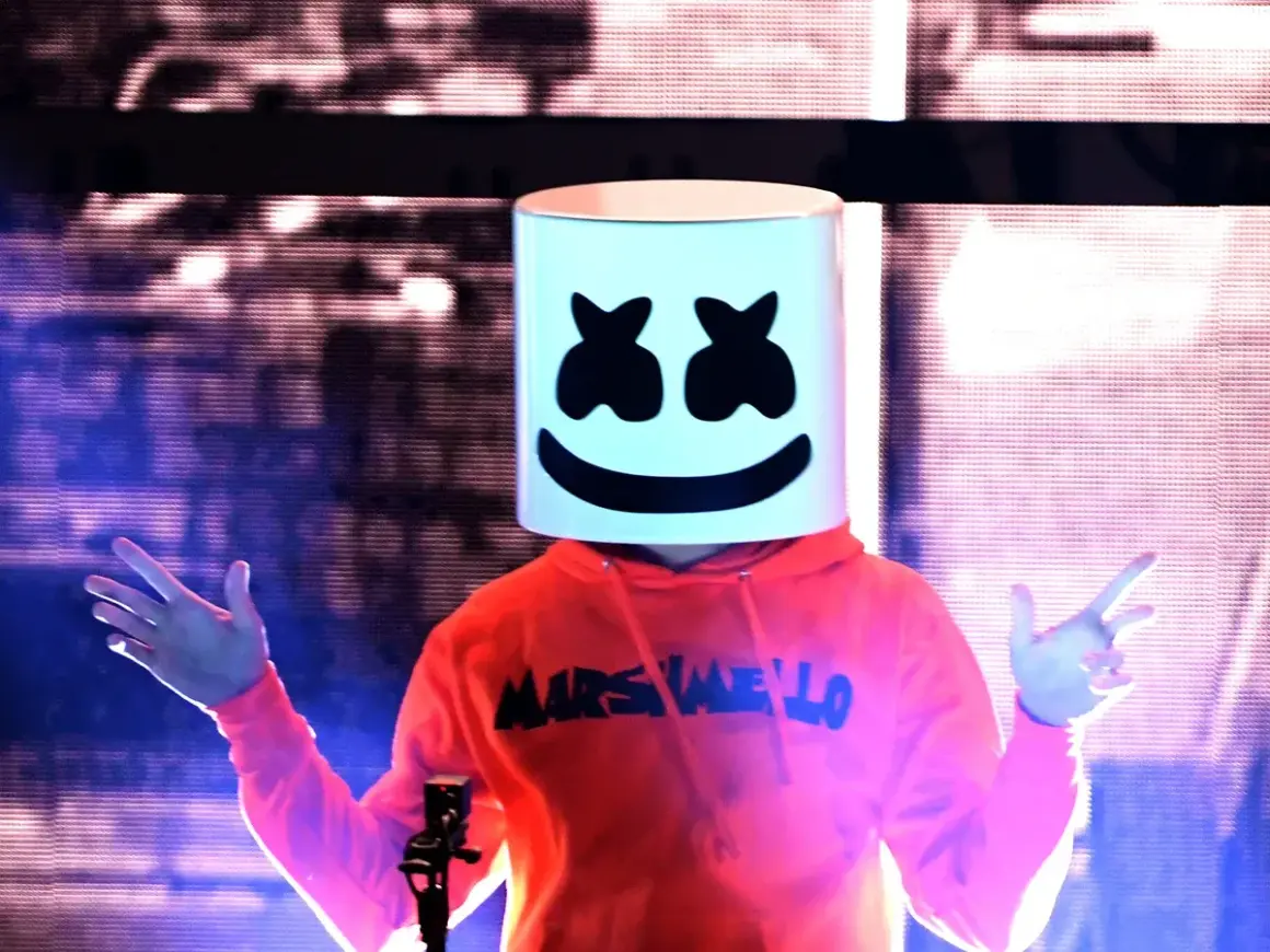 Christopher Comstock, conocido como Marshmello, con su icónica máscara blanca y sudadera naranja, saluda al público en un concierto.