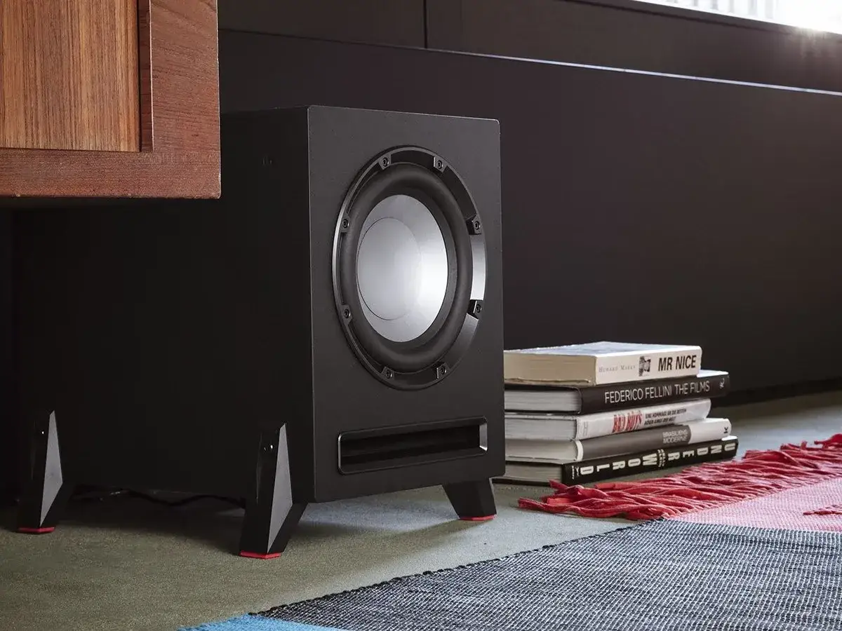 Subwoofer negro con patas rojas, junto a libros y una alfombra. Una idea de donde poner el subwoofer para un sonido envolvente.