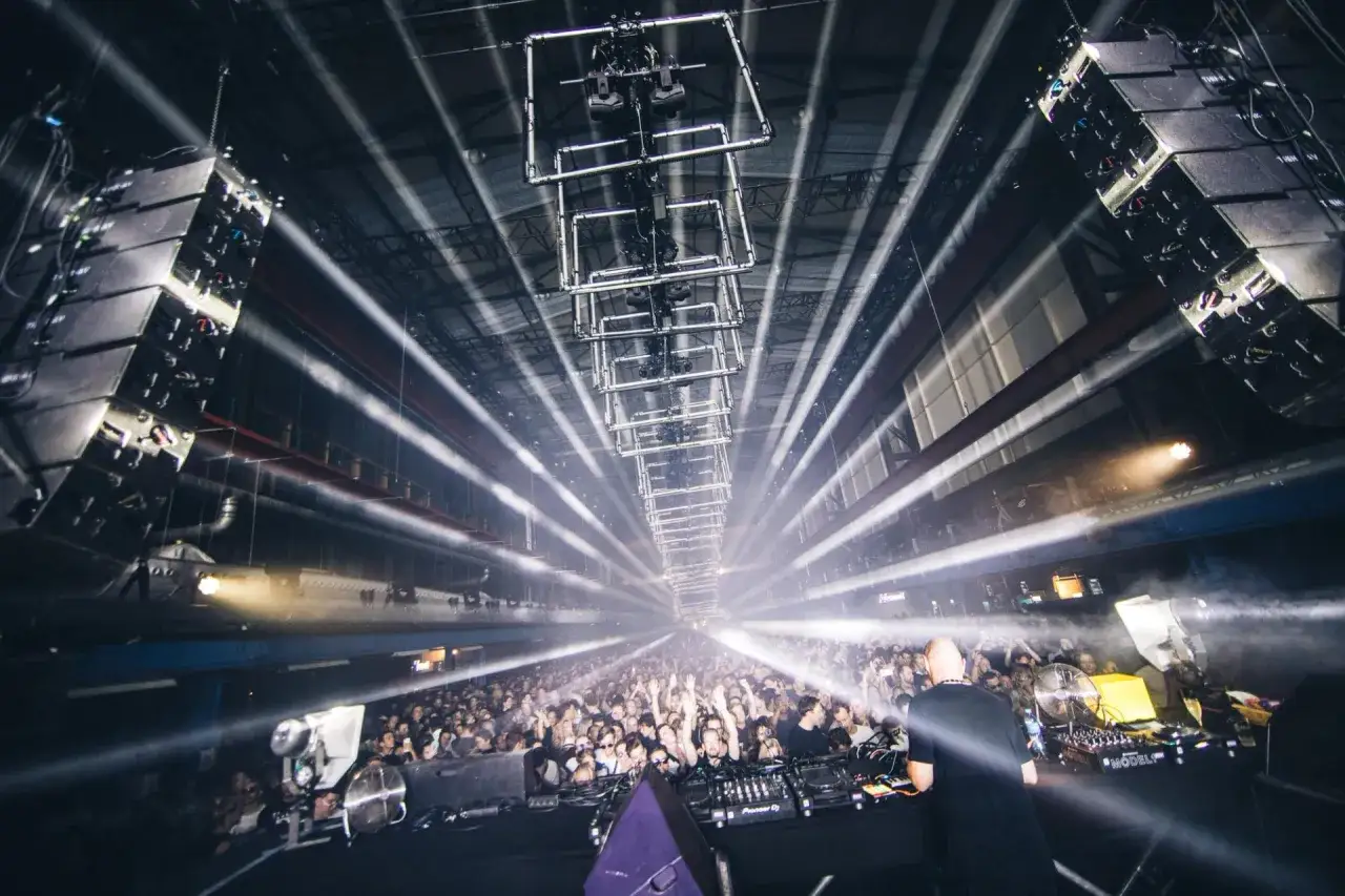 Un DJ pincha ante una multitud eufórica en un festival de música electrónica. Rayos de luz blanca atraviesan el humo, creando una atmósfera vibrante.