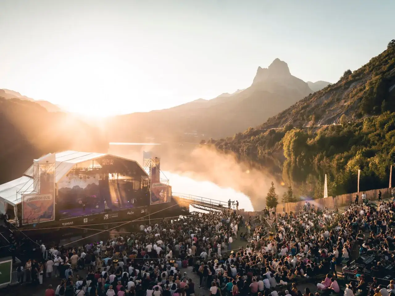 Gran multitud disfruta de un concierto al atardecer en uno de los festivales en octubre, con montañas y un lago de fondo.