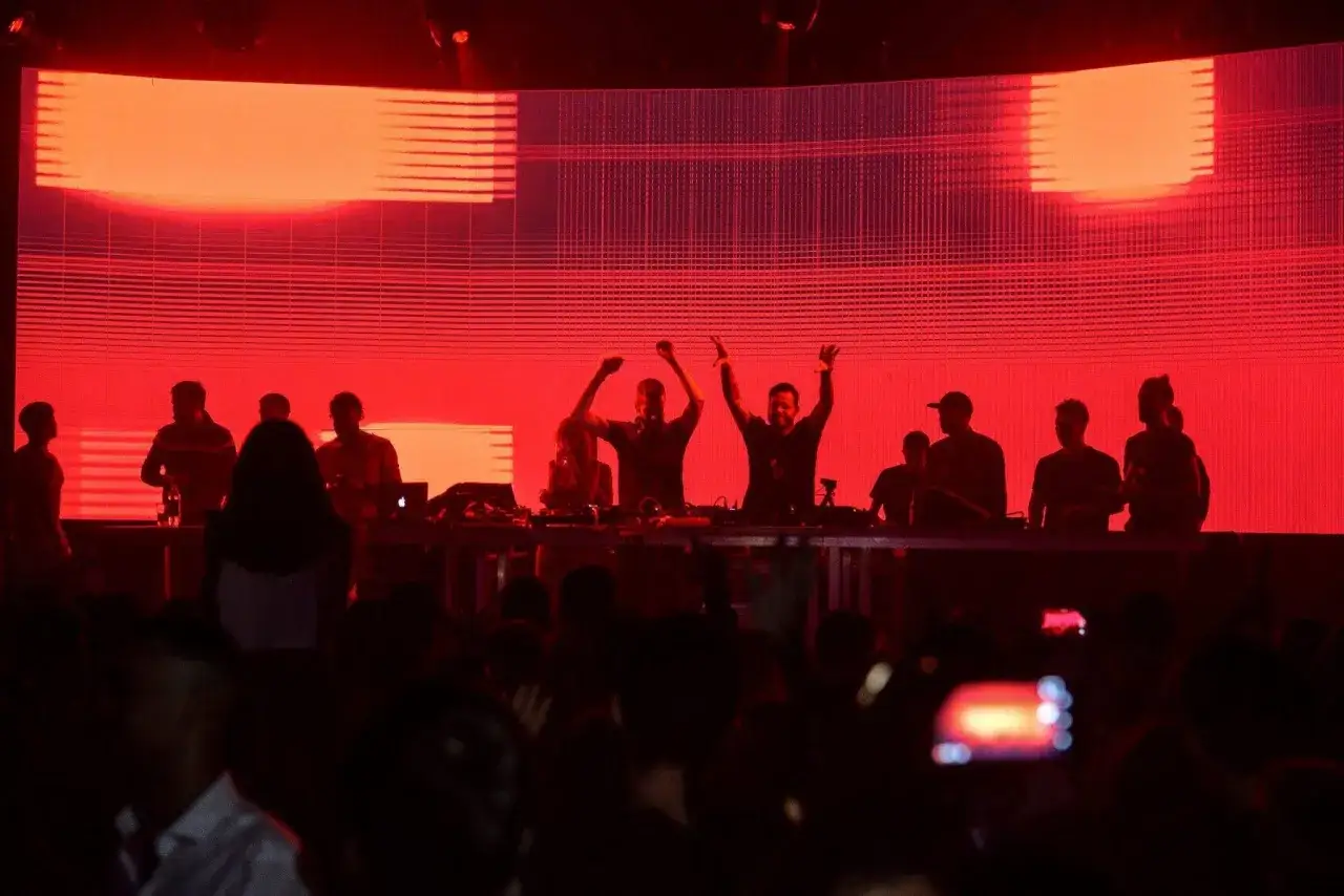 Un grupo de DJs en un escenario con luces rojas, animando a la multitud. As&iacute; se aprende como ser un buen DJ.