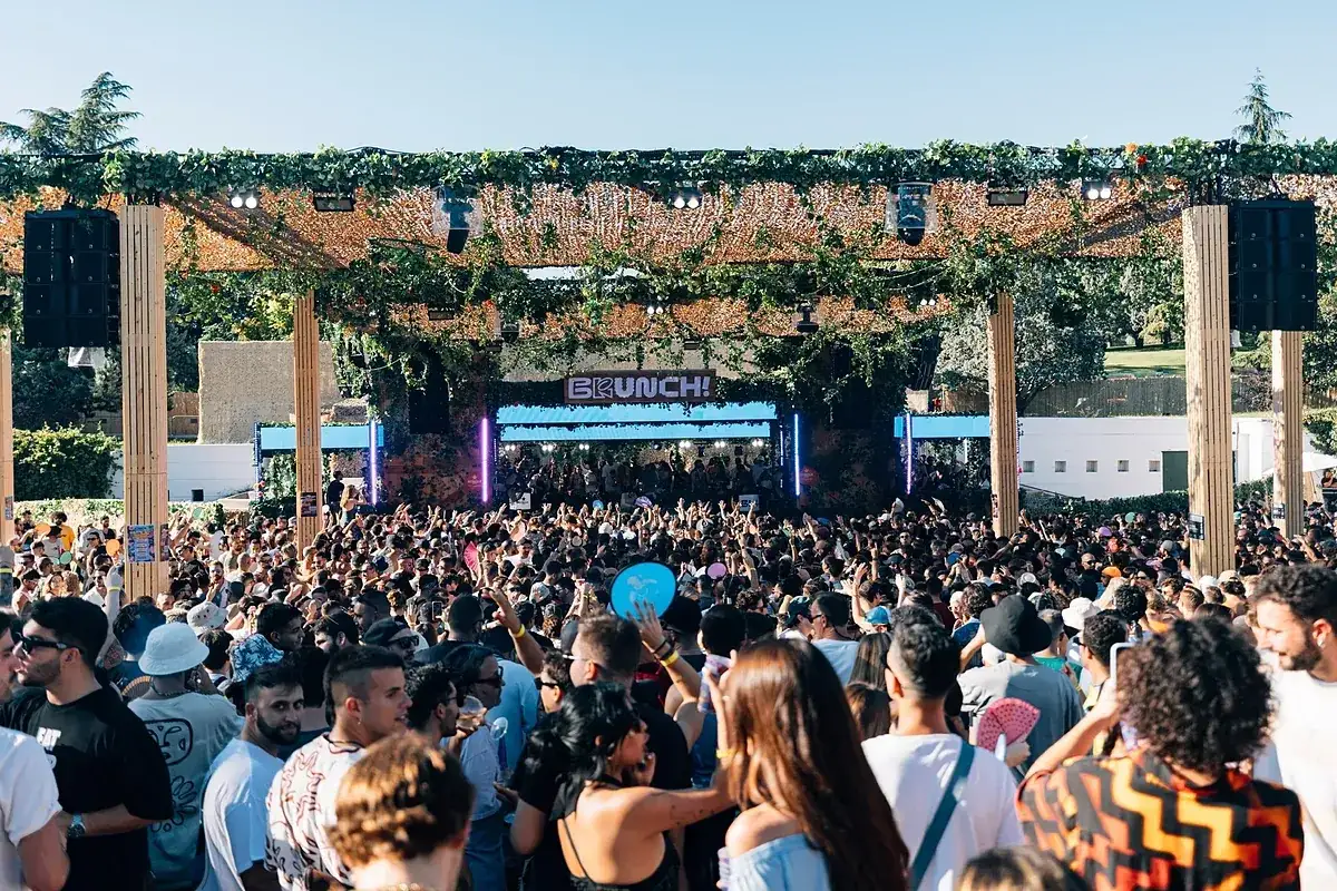Multitud disfrutando de un día soleado en Brunch Electronik Lisboa, con un escenario decorado con vegetación y luces vibrantes.