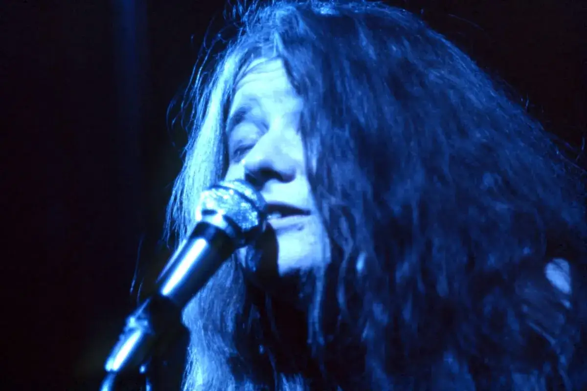 Janis Joplin, con su melena suelta y voz potente, canta apasionadamente al micr&oacute;fono bajo una luz azul.