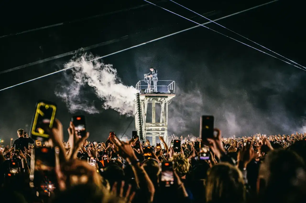 Un DJ pincha techno musica en una plataforma elevada, rodeado de humo y luces, mientras una multitud con m&oacute;viles levanta las manos en se&ntilde;al de &eacute;xtasis.