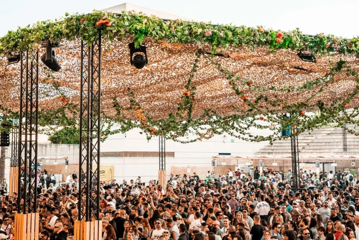 Multitud disfrutando de un evento al aire libre con una estructura decorada con vegetaci&oacute;n y luces, similar a un brunch electr&oacute;nico en M&aacute;laga.