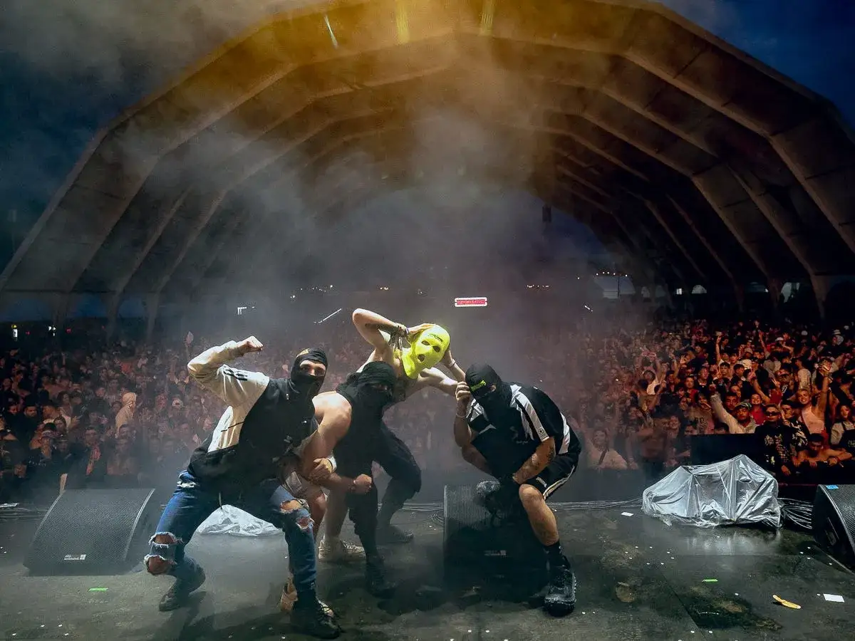 Artistas enmascarados act&uacute;an ante una multitud en un concierto. El p&uacute;blico parece vibrar con la energ&iacute;a, y los artistas, con sus pasamonta&ntilde;as y una m&aacute;scara amarilla, crean un ambiente de misterio y emoci&oacute;n.