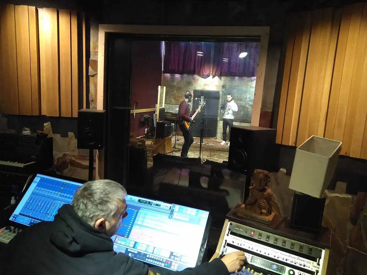 Un hombre trabaja en una mesa de mezclas mientras dos m&uacute;sicos ensayan en un estudio, un momento clave en su carrera musical.