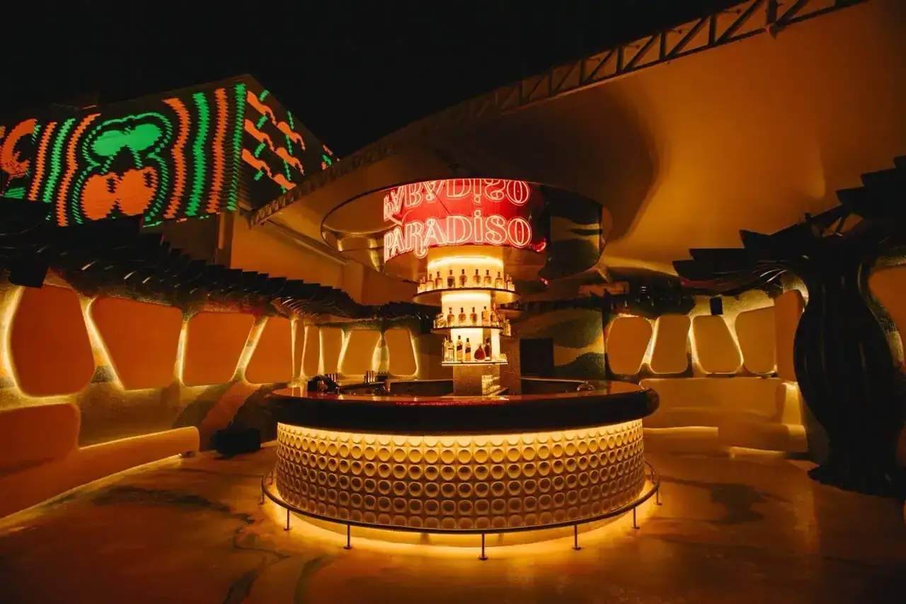 El interior de un club nocturno con un bar circular, luces de neón rojas que dicen