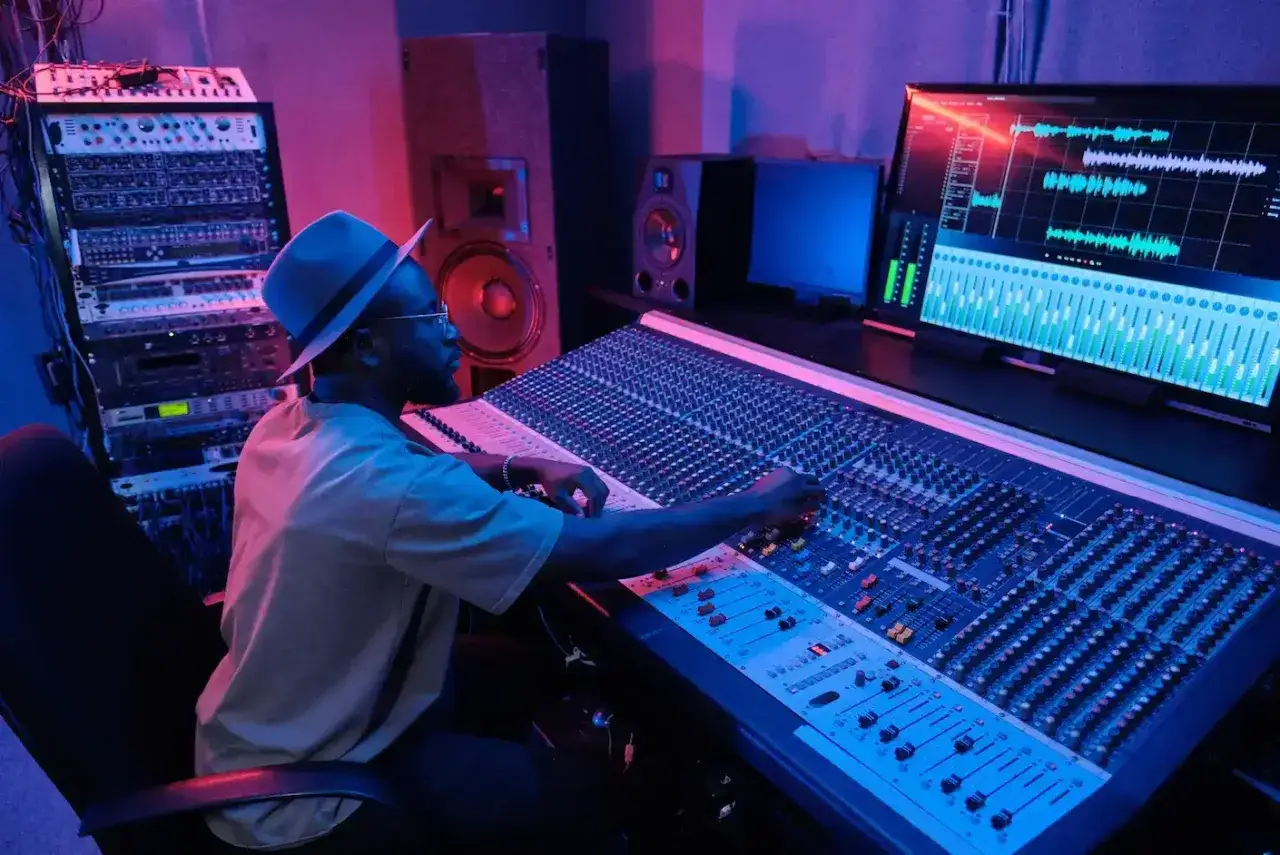 Productor musical ajustando controles en una mesa de mezclas, rodeado de equipos de audio y luces de neón.