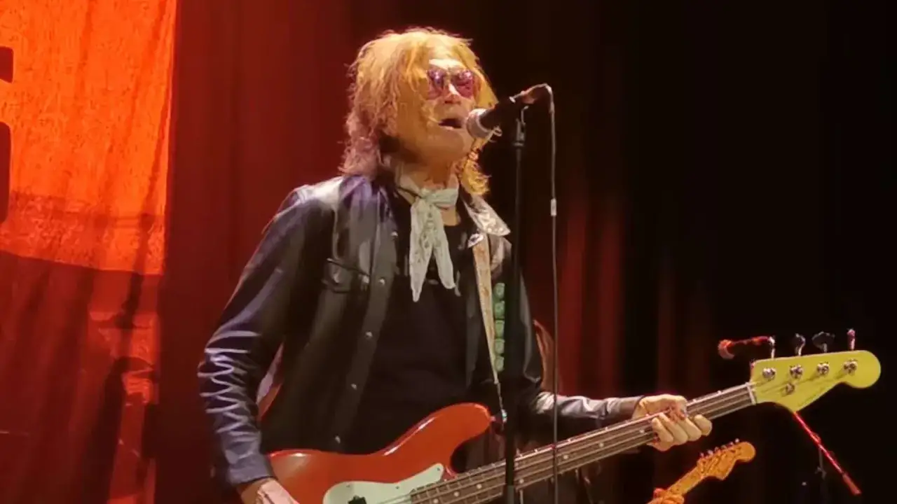 Glenn Hughes, con su banda, canta y toca el bajo en un escenario con cortinas rojas.