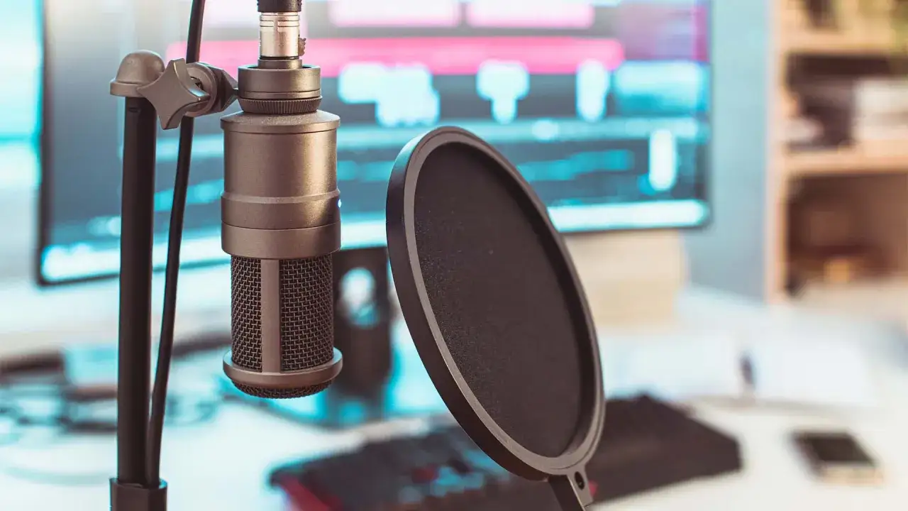 Un micrófono de presión listo para grabar en un estudio casero, con una pantalla anti-pop y un ordenador con software de edición de audio al fondo.
