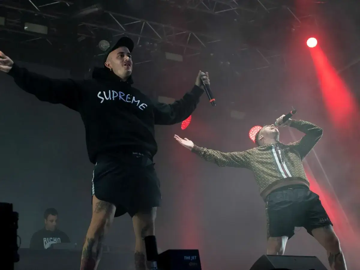Dos artistas de hip hop español cantan en un escenario con luces rojas. Uno lleva una sudadera negra