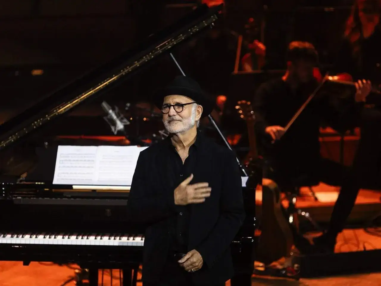 Ludovico Einaudi, con su caracter&iacute;stica edad y sombrero, dirige una orquesta junto a un piano.