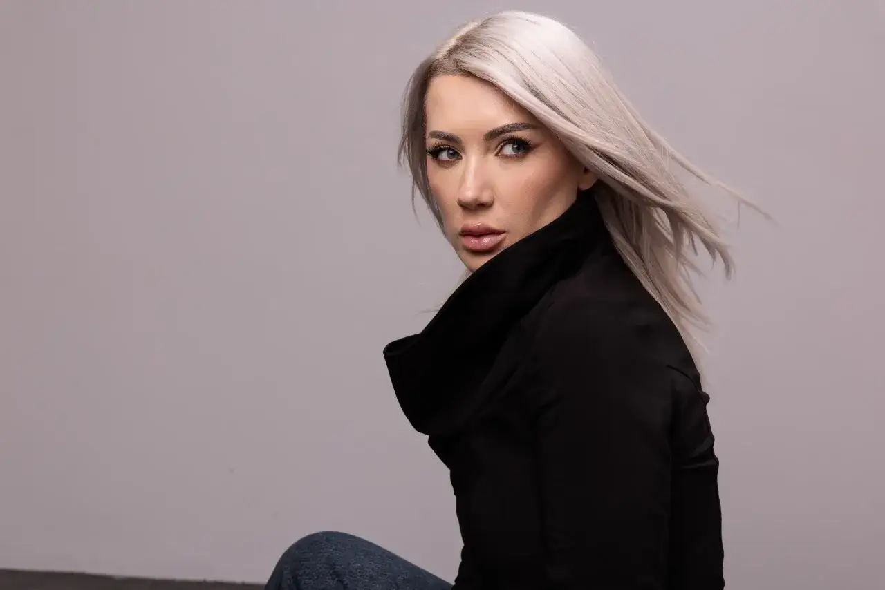 Nusha, con su cabello rubio platino y mirada intensa, posa con un elegante abrigo negro.
