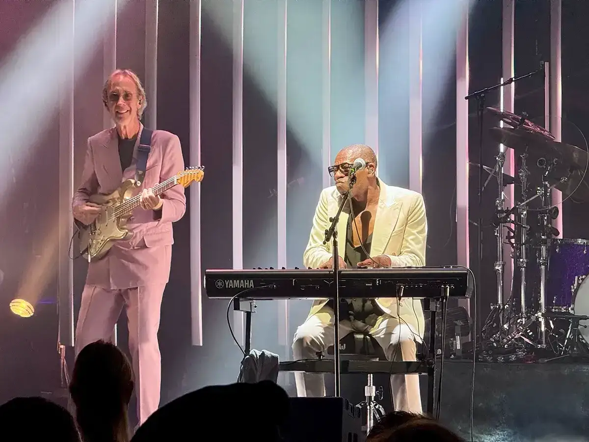 Miembros de Mike and the Mechanics en concierto: un guitarrista sonriente con traje rosa y un teclista con traje blanco.