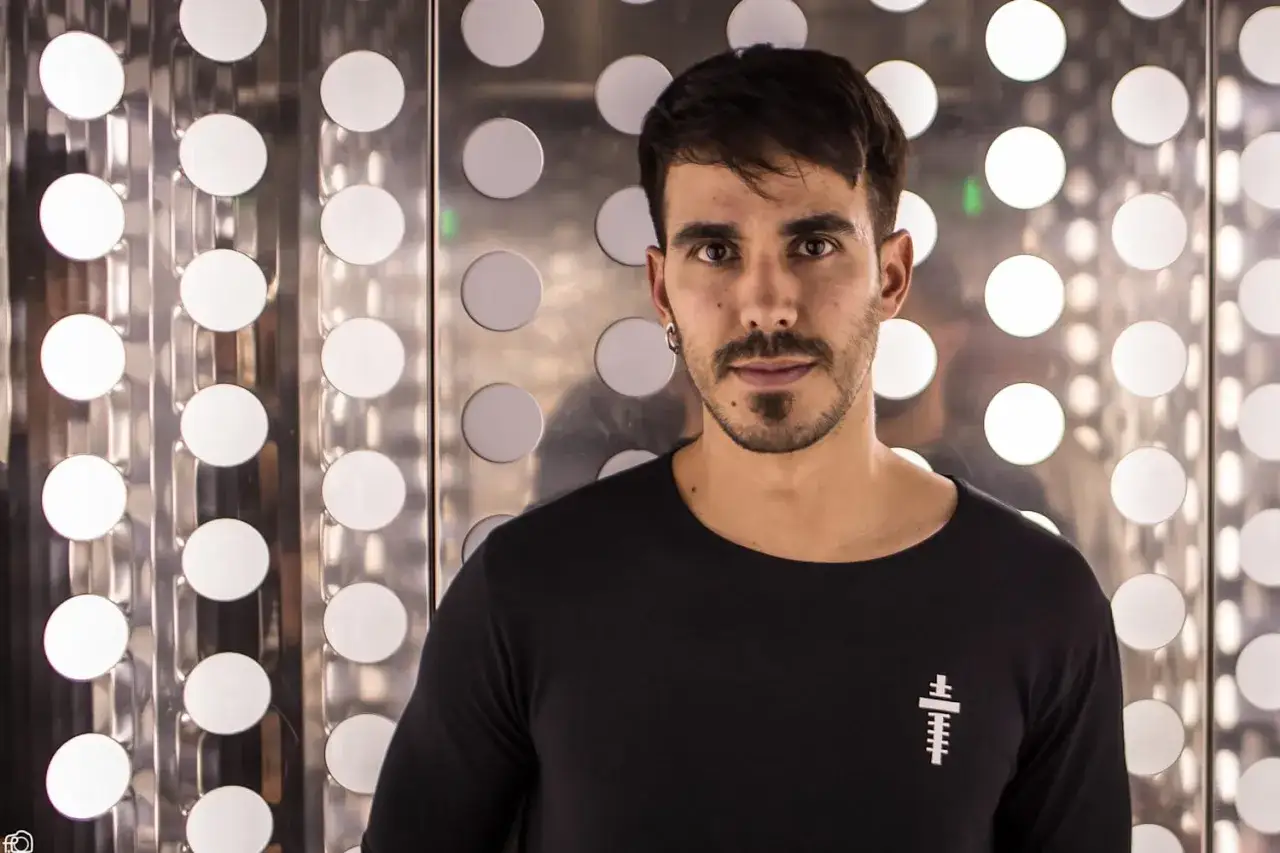 Raúl Pacheco DJ, con su mirada intensa, posa frente a un fondo metálico con círculos luminosos.
