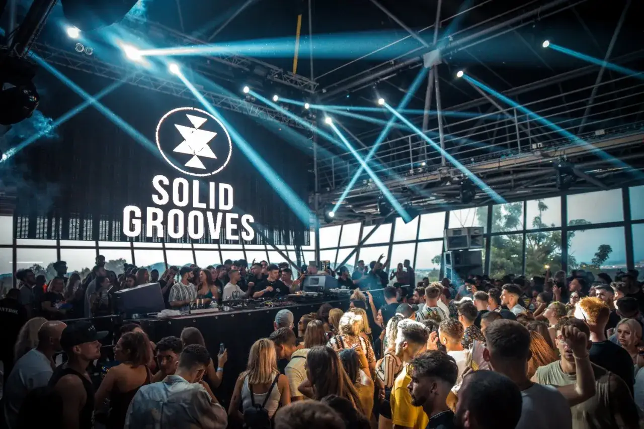Multitud bailando bajo luces estrobosc&oacute;picas en un club con el logo de Solid Grooves Ibiza en la pantalla.