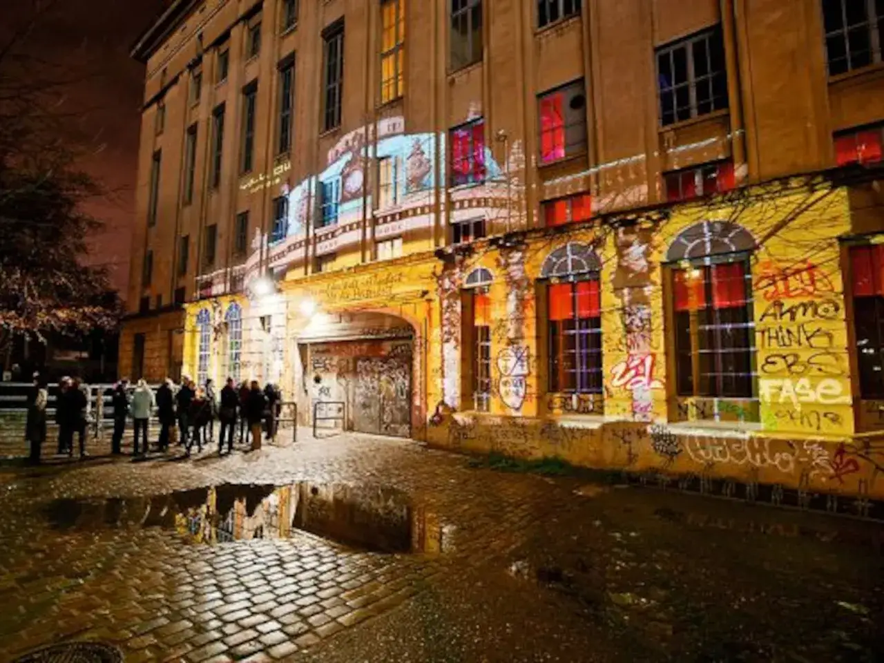 Fachada de edificio con grafitis y proyecciones de luces, como un berghain simulator. Reflejos en charcos en el suelo adoquinado.
