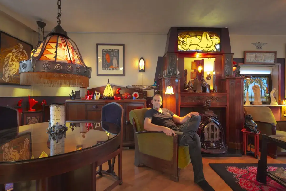 Hombre sentado en sillón en una sala de estilo Art Nouveau, con lámparas y muebles de madera oscura. Un detalle de