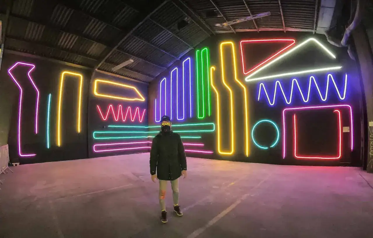 Persona con gorra y bufanda frente a un muro con luces de ne&oacute;n de colores. Inspiraci&oacute;n para nombres para cantantes de trap.